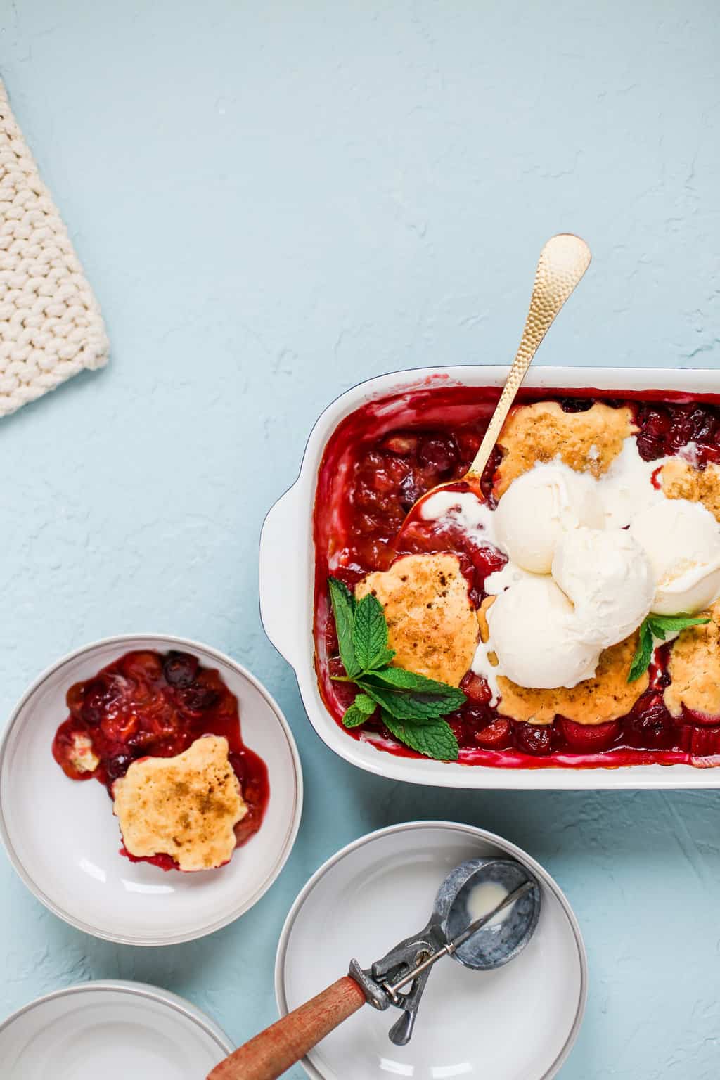 a casserole dish of cherry rhubarb cobbler with a serving in an individual bowl beside it, and the ice cream scooper resting in a separate bowl