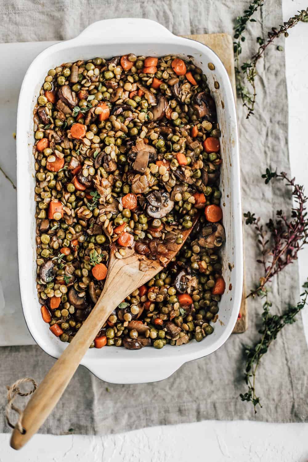 Casserole dish filled with sautéed carrots, mushroom, peas, onions, and lentils and a wooden spoon.