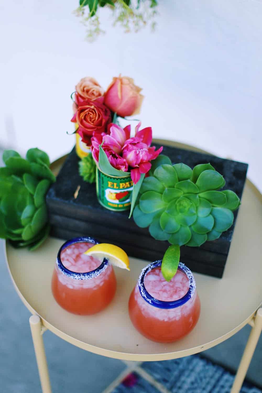 Two margaritas on the rocks with flowers in Mexican food cans on a beige table