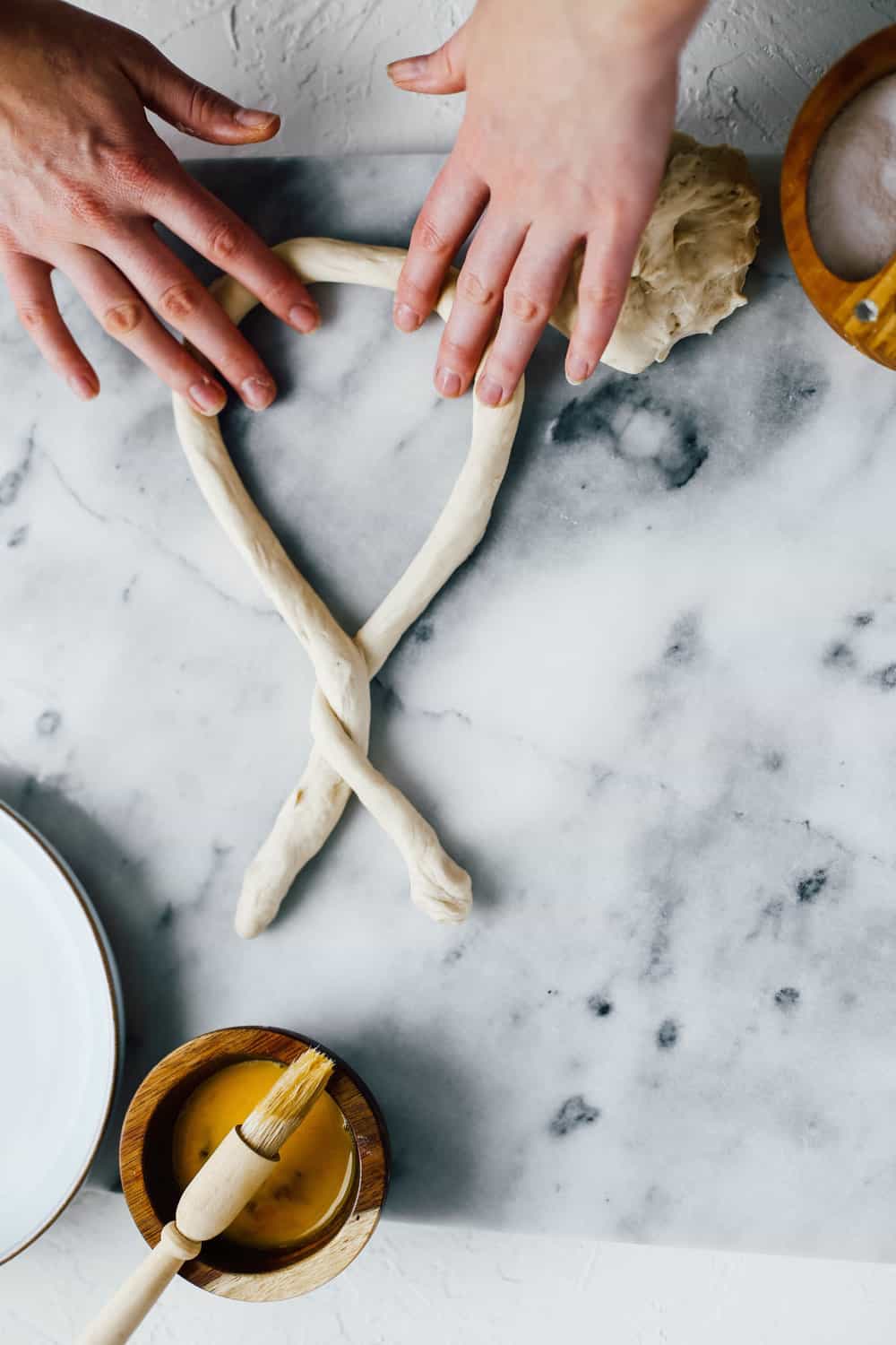 two hands twisting pretzel dough on marble countertop