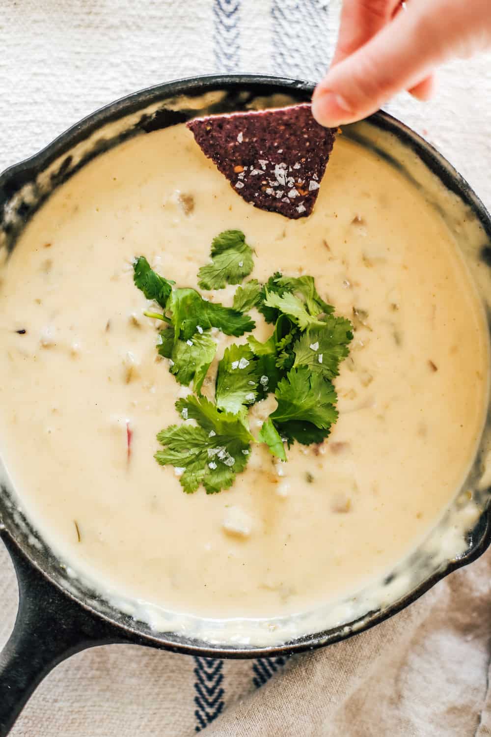 A skillet of green chile queso topped with cilantro and a chip being dipped. 