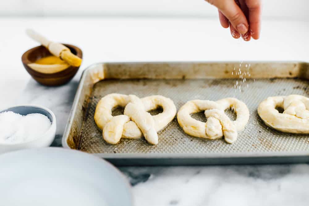 hand sprinkling salt on two shaped pretzels on baking sheet