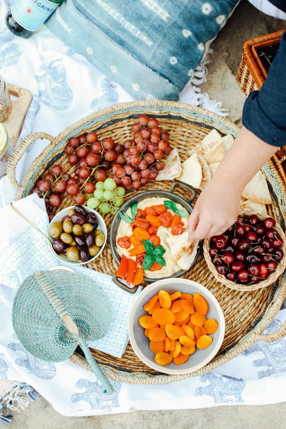 Hand dipping pita in a bowl of hummus in a wicker basket with grapes cherries apricots and olives on beach towels