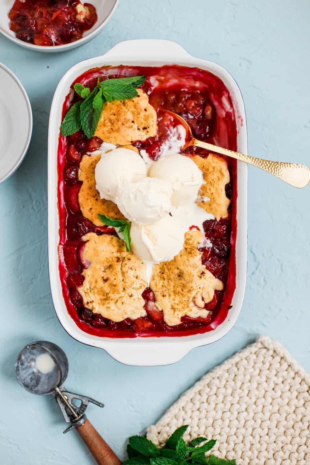 a casserole dish full of cherry rhubarb cobbler topped with vanilla ice cream and a large serving spoon sticking out of it 