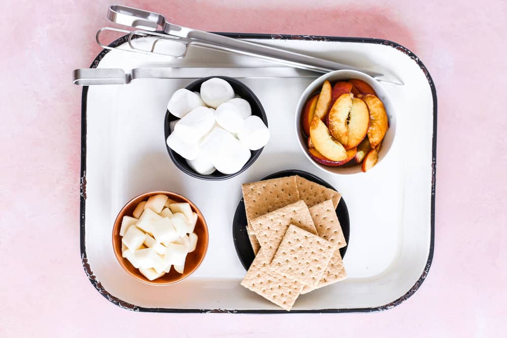 Overhead of prepped marshmallows, peaches, white chocolate, and graham crackers with metal skewers