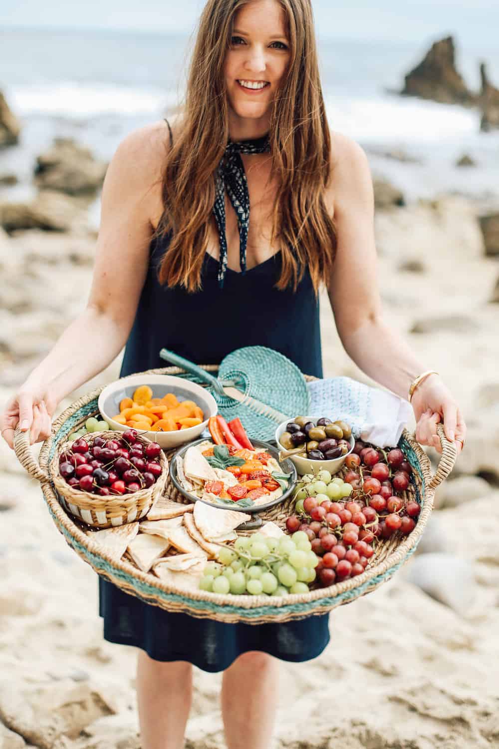 Elizabeth holding a wicker basket full of grapes pita cherries apricots olives and hummus on the beach