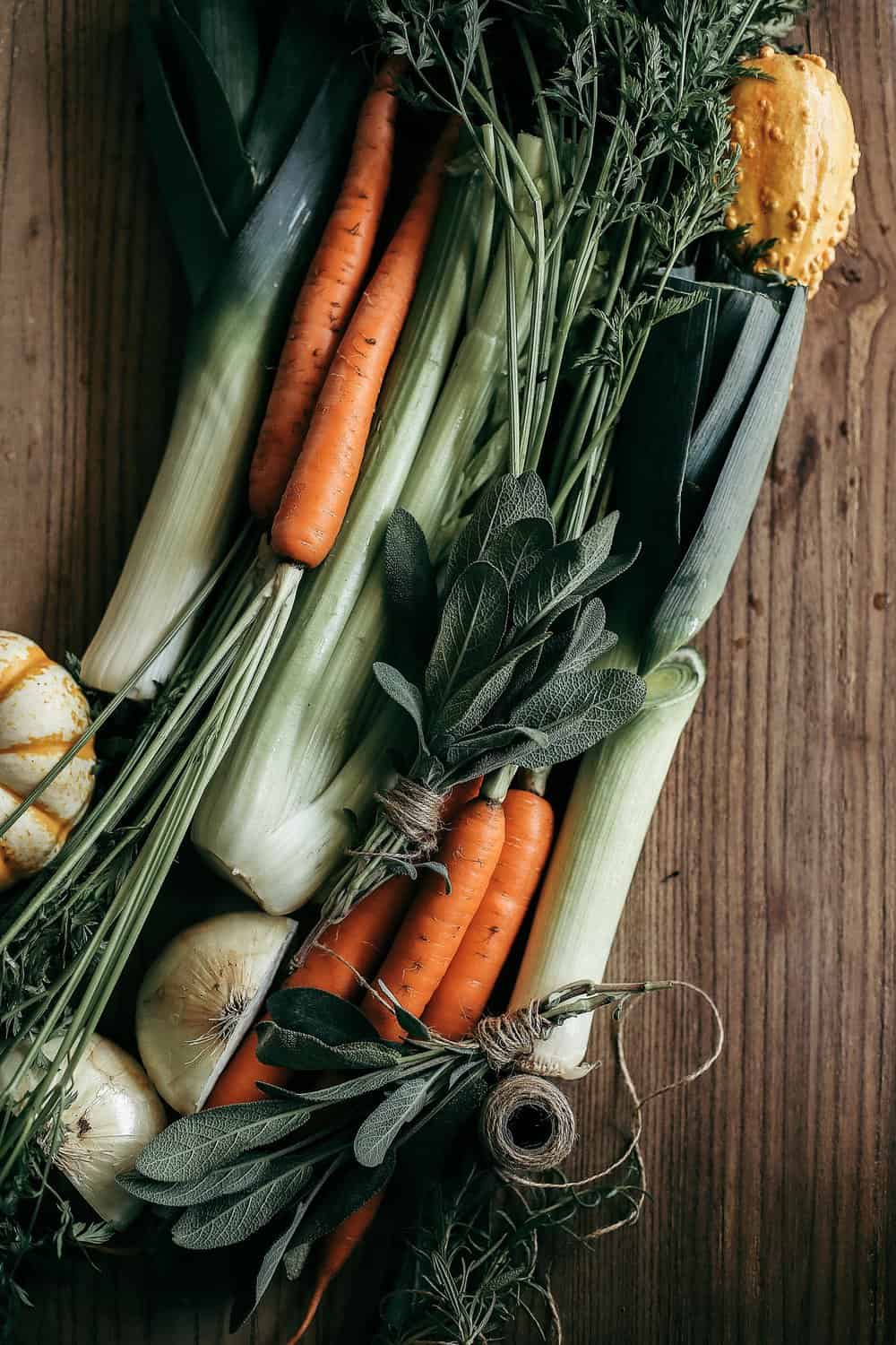 Celery, carrots, leeks, onion, and sage on a wooden cutting board. 