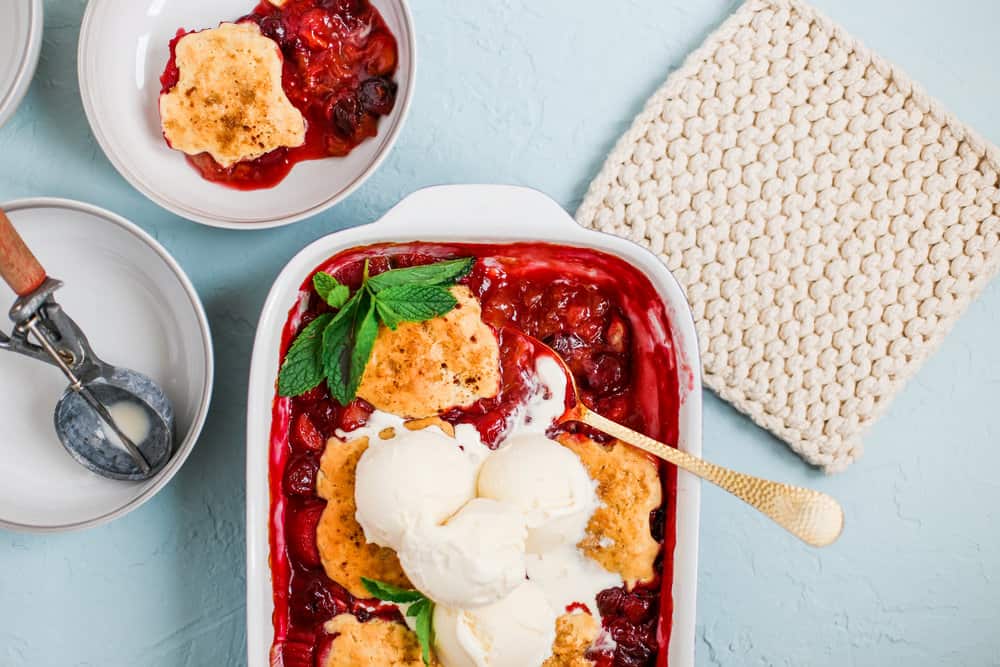 the cherry cobbler in a casserole dish beside a plate full of it, the ice cream scooper, and a square of woven cloth for a hot dish