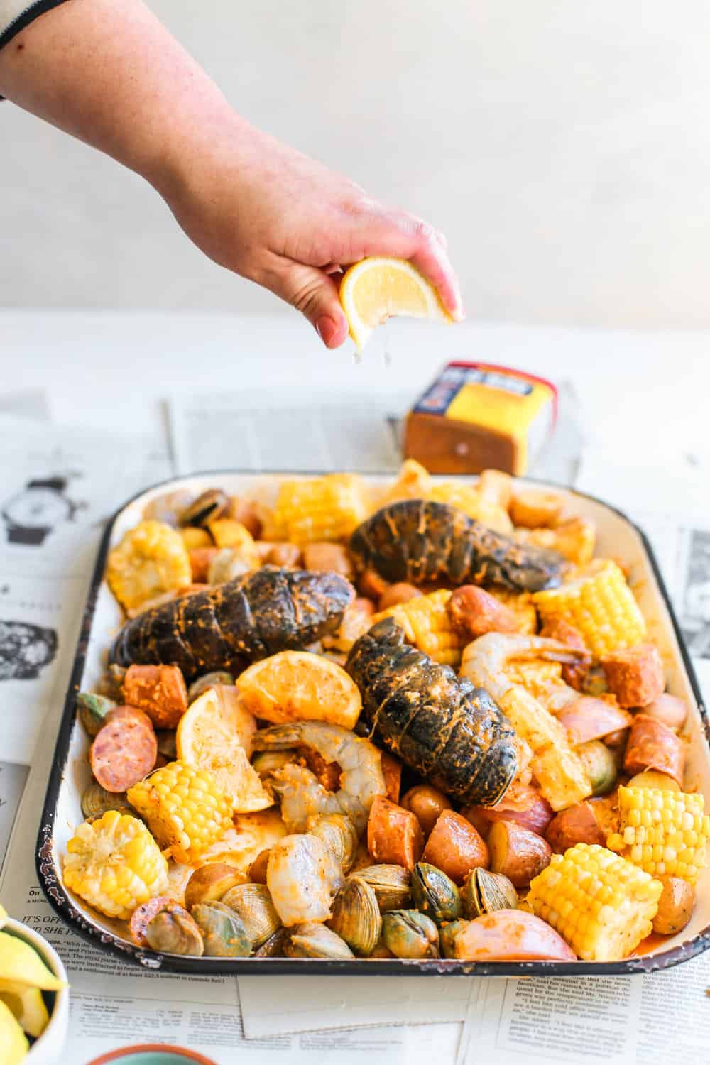 hand squeezing lemon over baking sheet with lobster, corn, potatoes