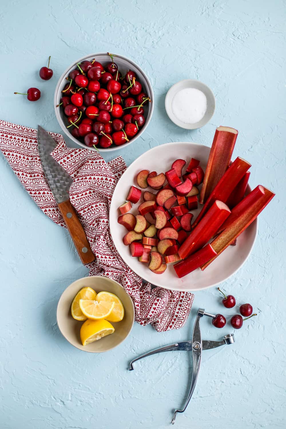 rhubarb, cherries, sugar, lemon, a pitter, and a knife on a light blue surface