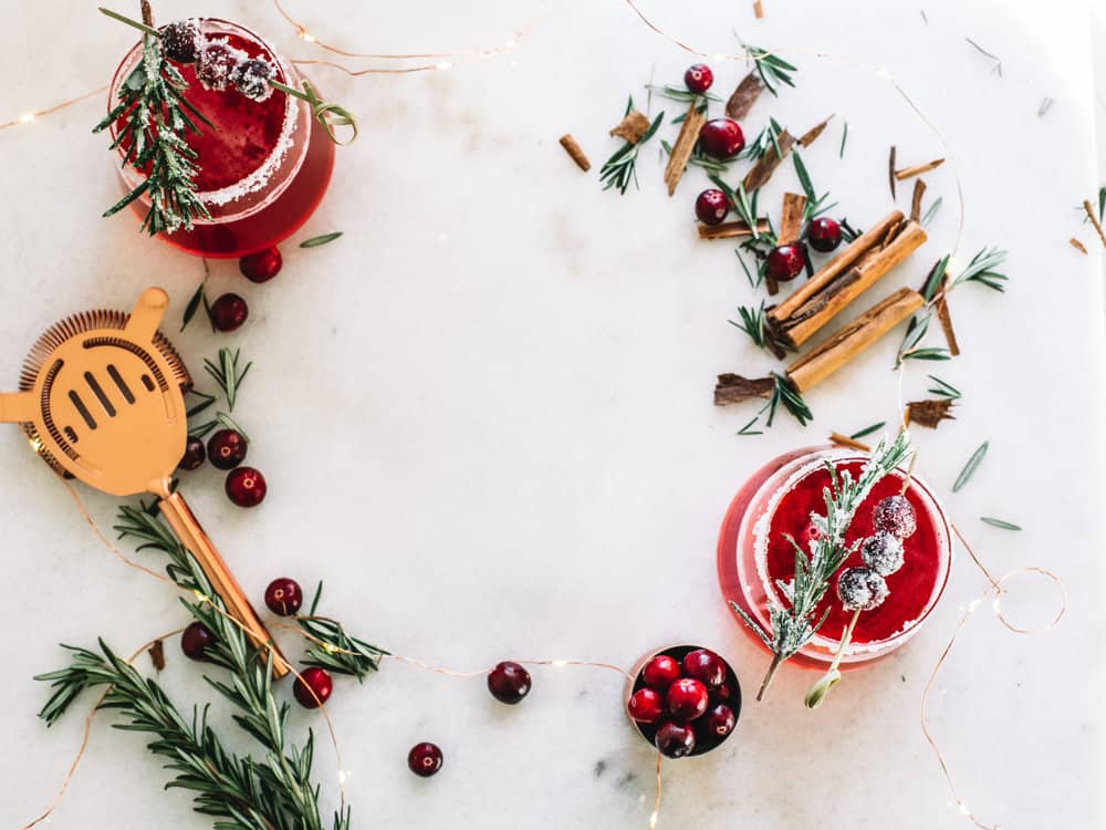 Low glasses filled with a cranberry whiskey cocktails garnished with frosted cranberries and a rosemary sprig. With cinnamon sticks and a copper strainer. 