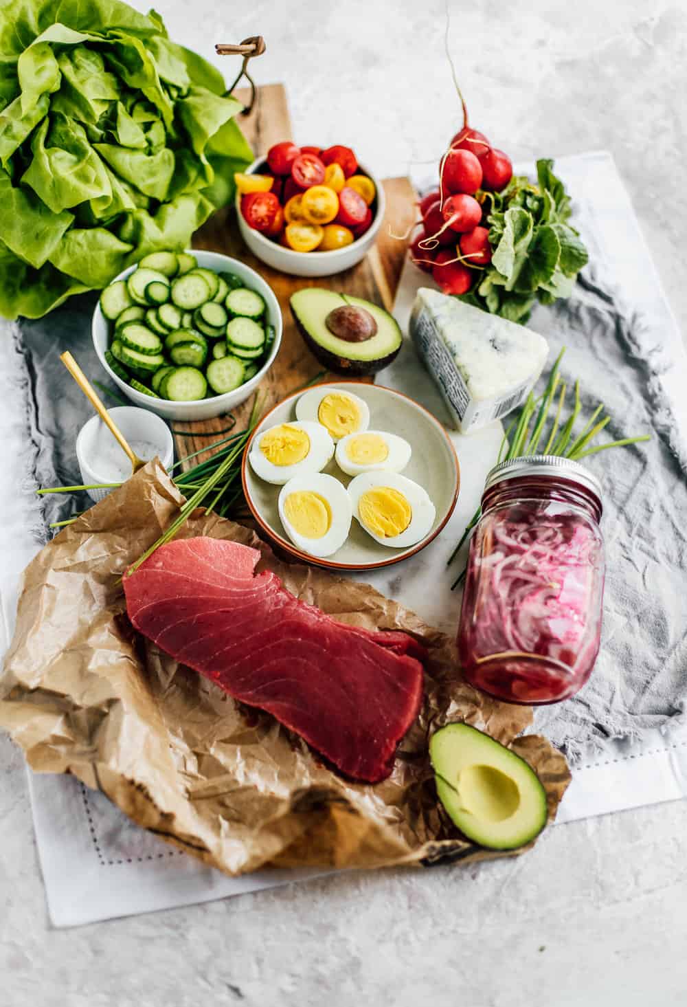 hard boiled eggs, a jar of pickled onions, radish, ahi tuna steak, avocado, cucumbers, tomatoes, blue cheese, and butter lettuce laid out n a board