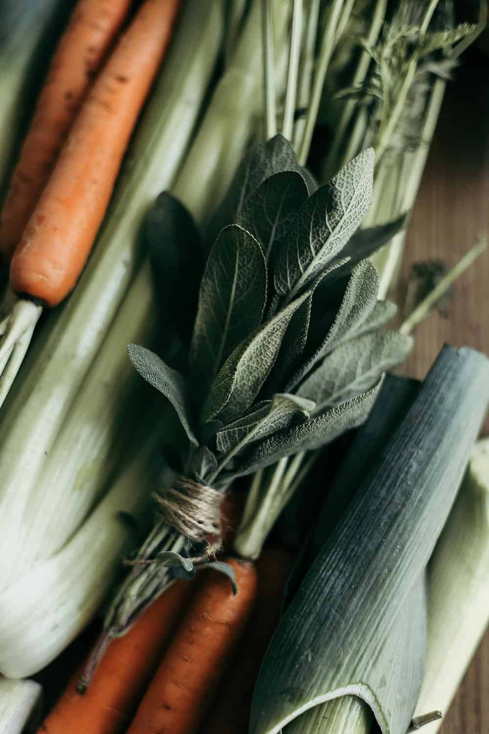 Leeks, carrots, celery, and bundled sage on a wooden cutting board.