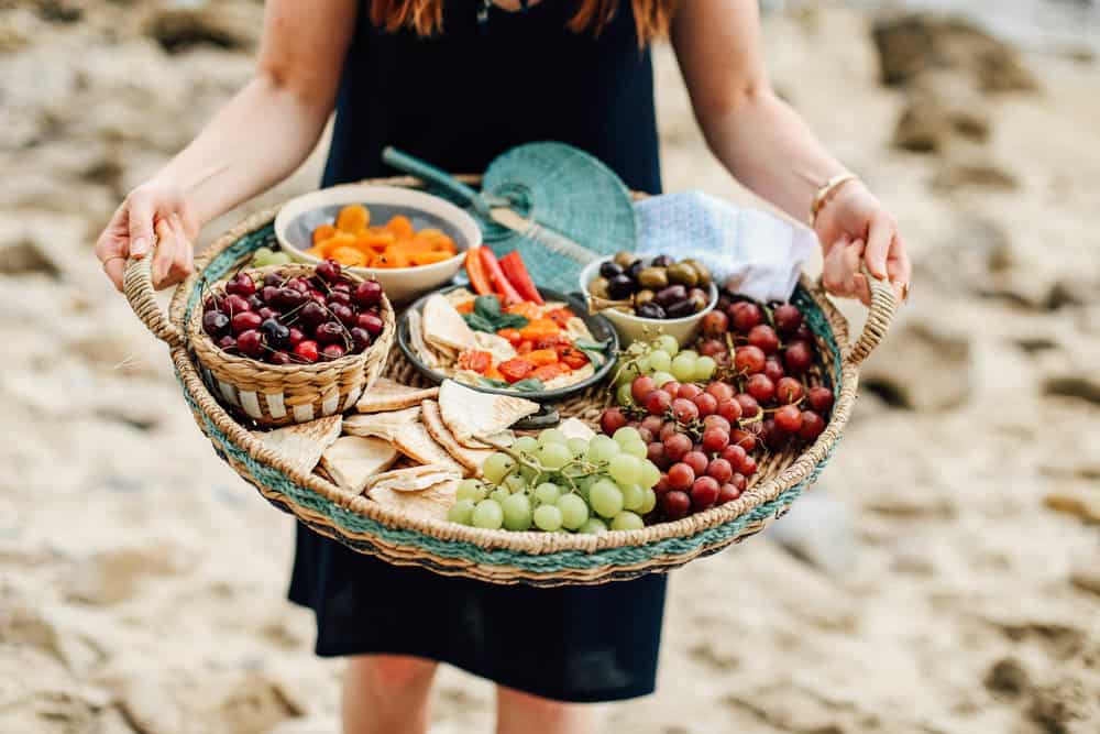 Elizabeth holding a basket full of grapes pita cherries apricots hummus and olives on the beach