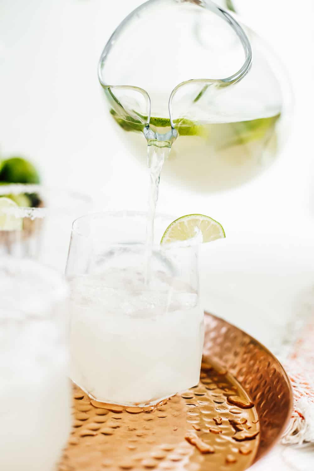 A low clear glass, being filled by a pitcher of a classic margarita on a copper tray. 