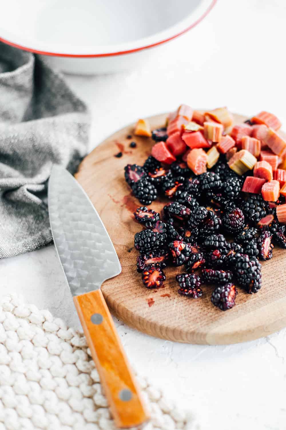 an oval brown cutting board with blackberries and rhubarb chunks on it beside a chef's knife