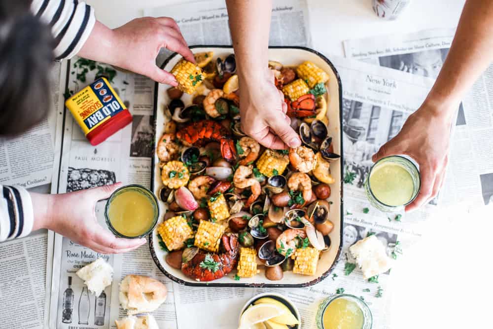 hands touching clams, shrimp, lobster, potatoes, corn on sheet pan with old bay and melted butter