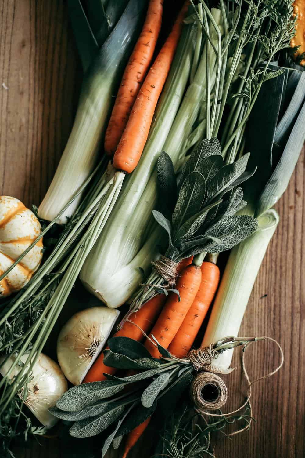 Carrots, celery, onions, leeks, bundled sage on a wooden cutting board. 