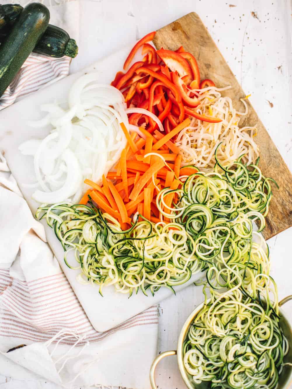 A cutting board with sliced onions, carrots, bell pepper, bean sprouts, and zucchini noodles.