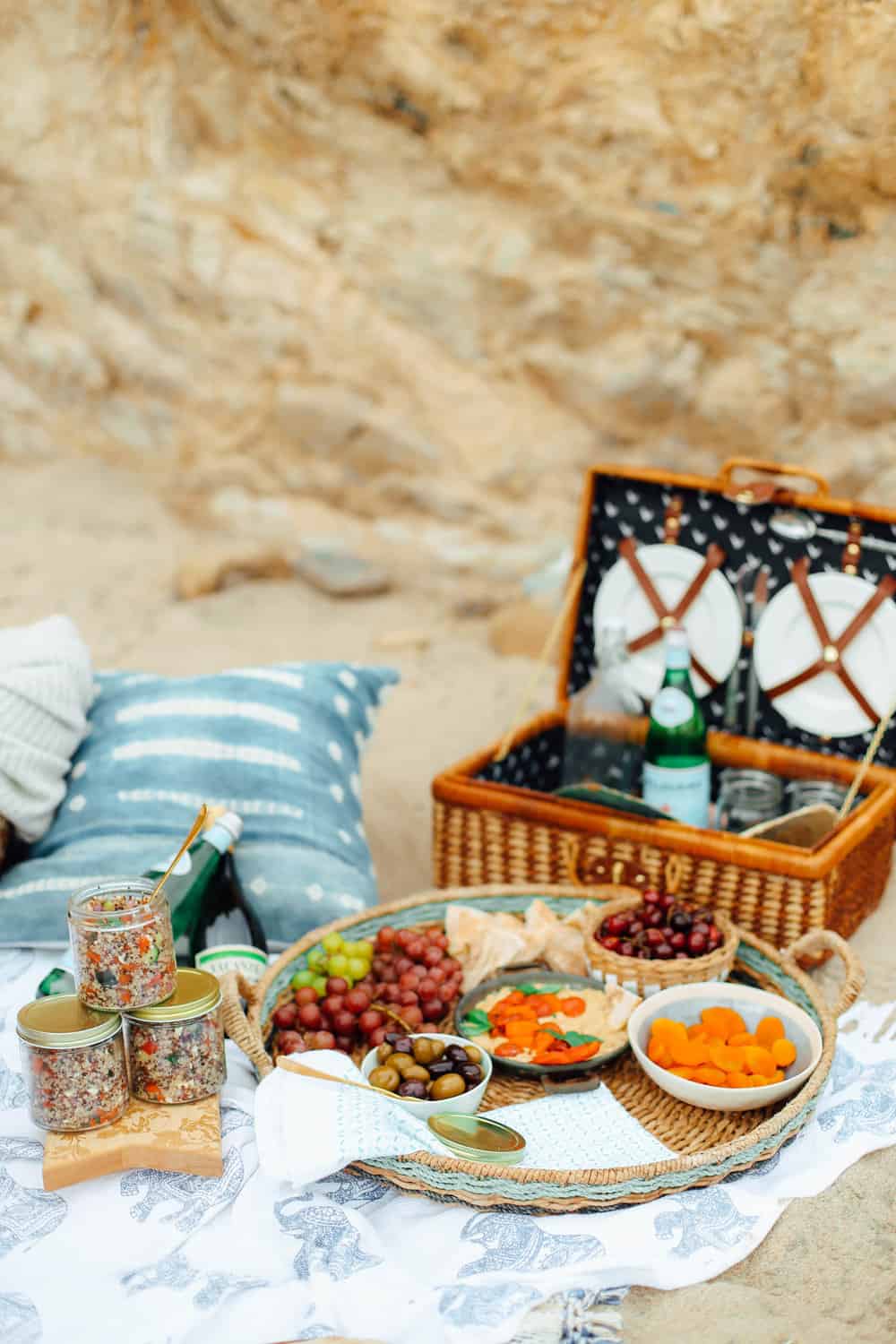 Grapes Pita Cherries Apricots Olives and Hummus in a wicker basket and sparkling water glasses and plates in a picnic basket with towels on the beach