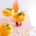 three glasses full of strawberry and peach frose in front of a bottle of rosé with ice cubes scattered around
