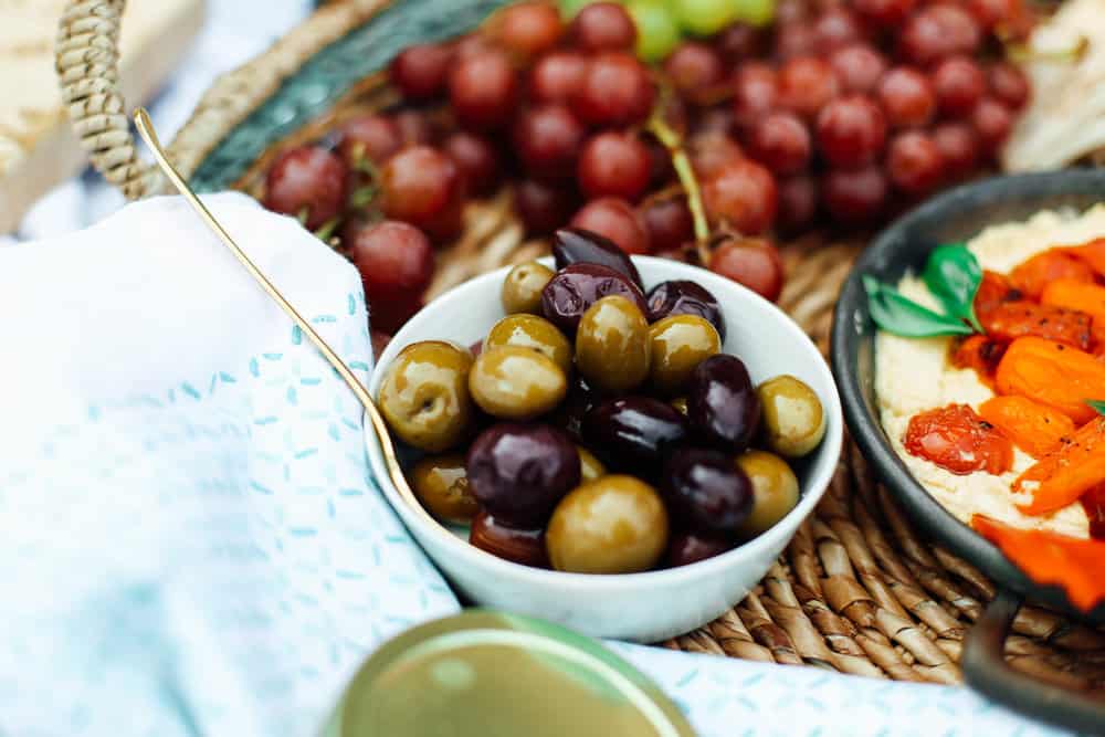 Mediterranean Olives in a white bowl with grapes and hummus in a wicker basket