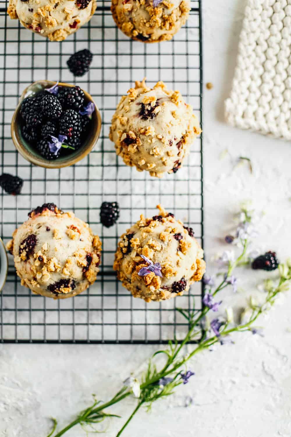 an overhead shot of blackberry and rhubarb muffins on the black cooling rack, beside a small bowl of blackberries and a sprig of flowers