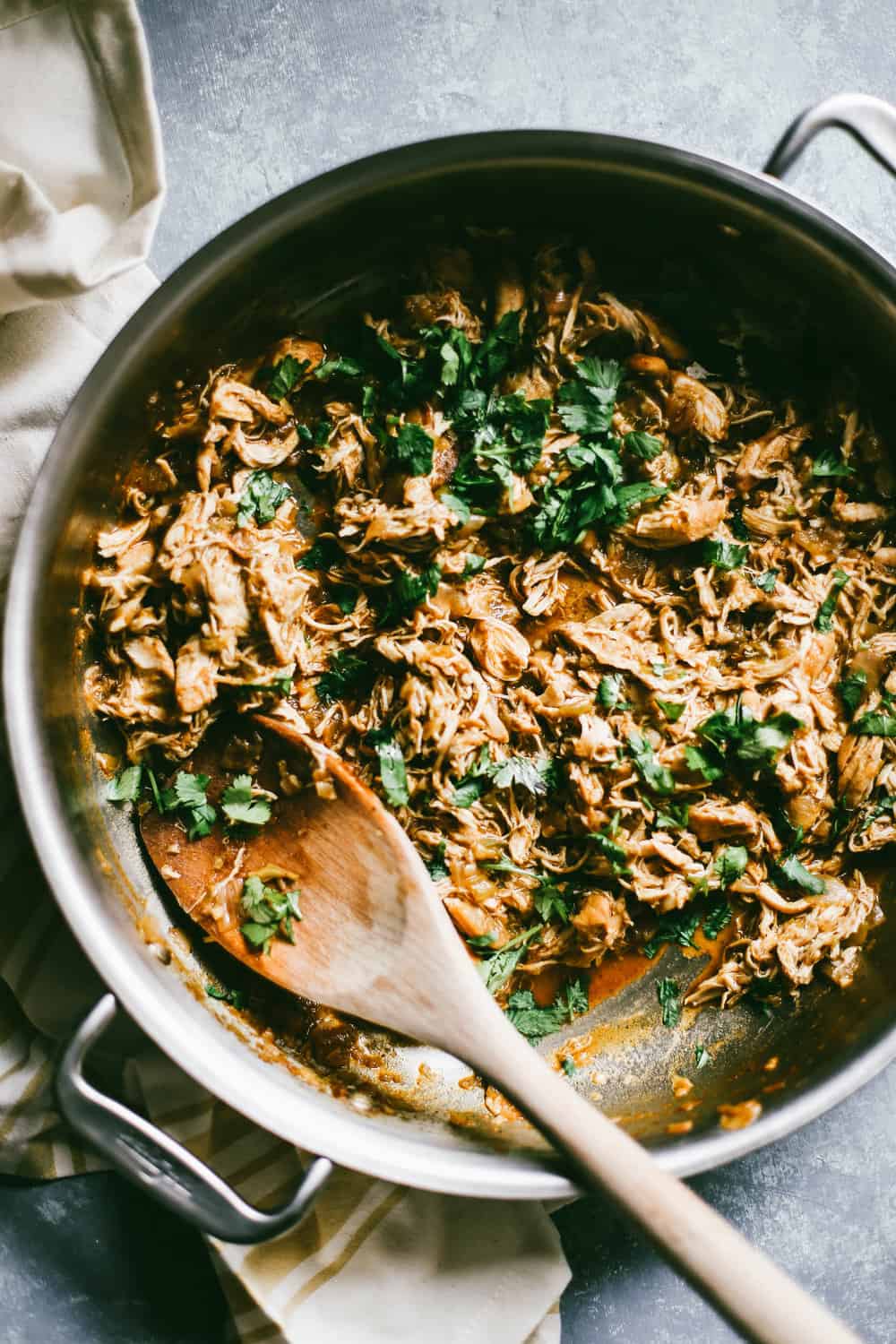 A pan with shredded chicken and cilantro being stirred with a wooden spoon. 