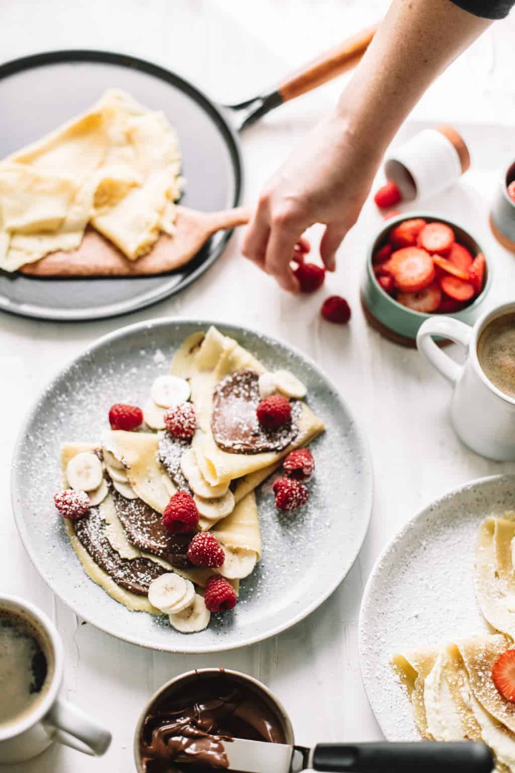 A plate full of crepes with sliced bananas, raspberries, and Nutella.