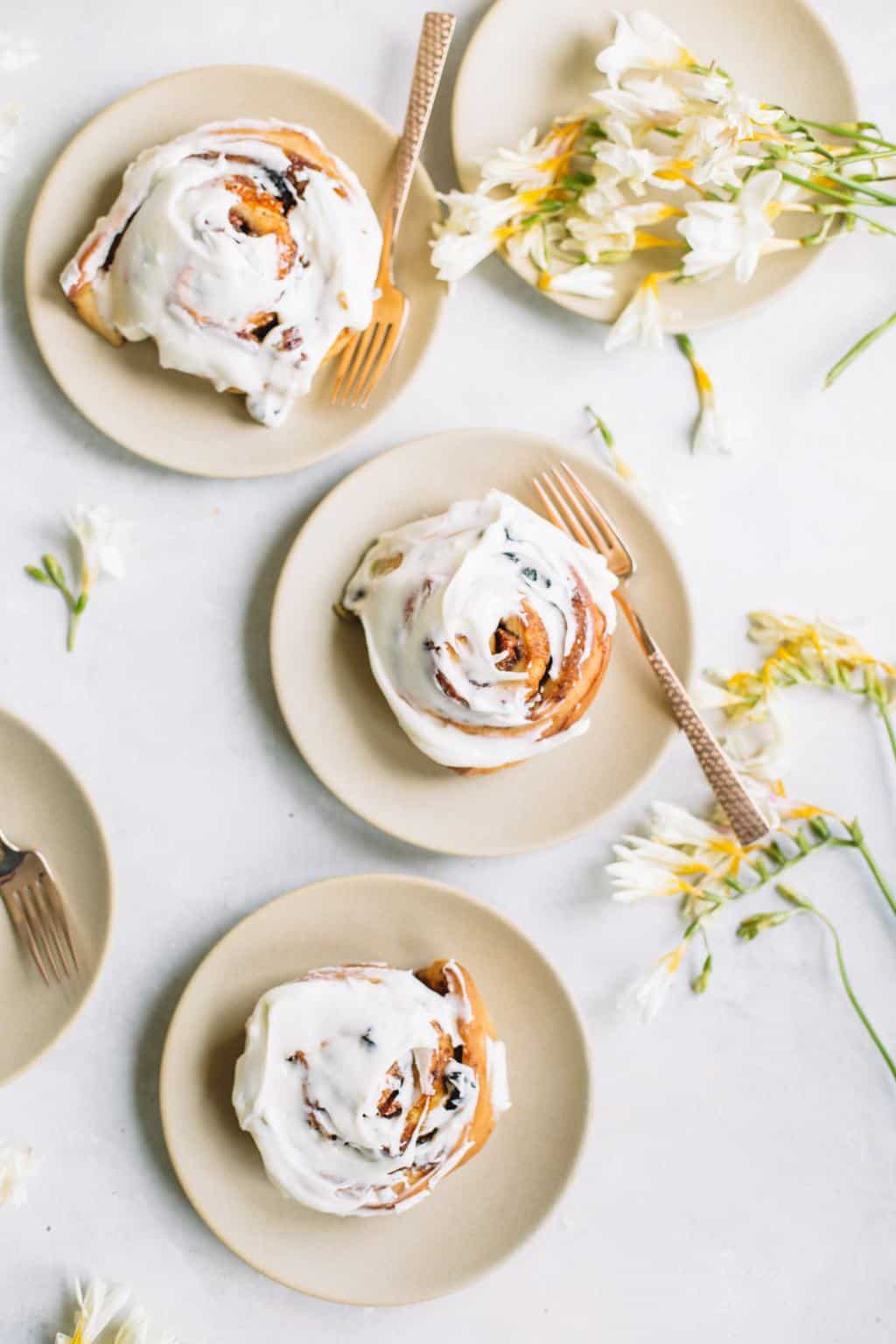 three cinnamon rolls with cream cheese icing on beige plate with gold fork and flowers