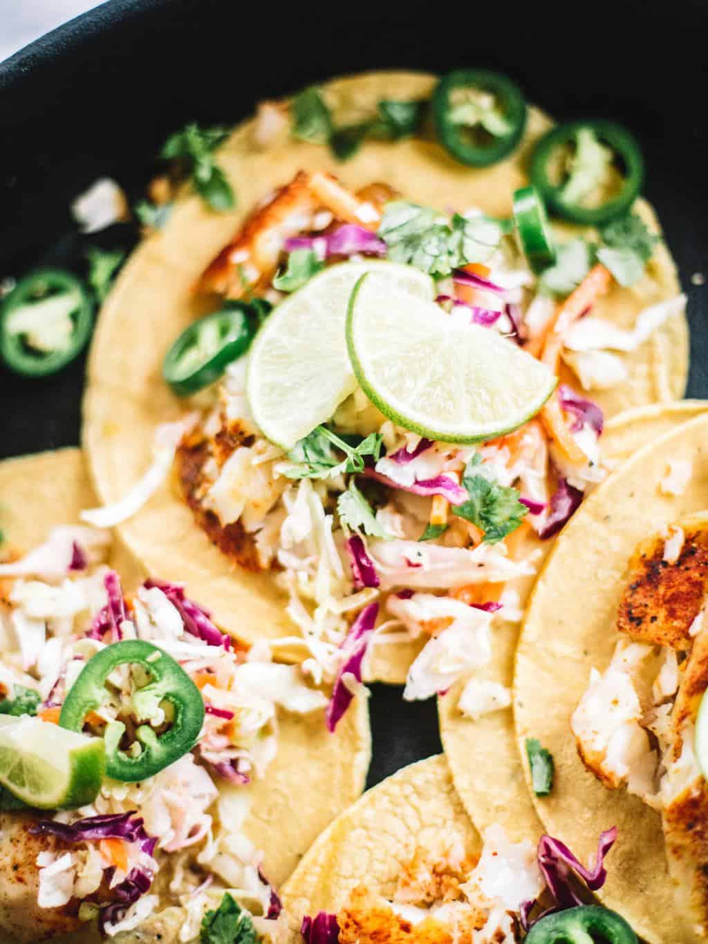 A black platter of fish tacos on corn tortillas with limes.