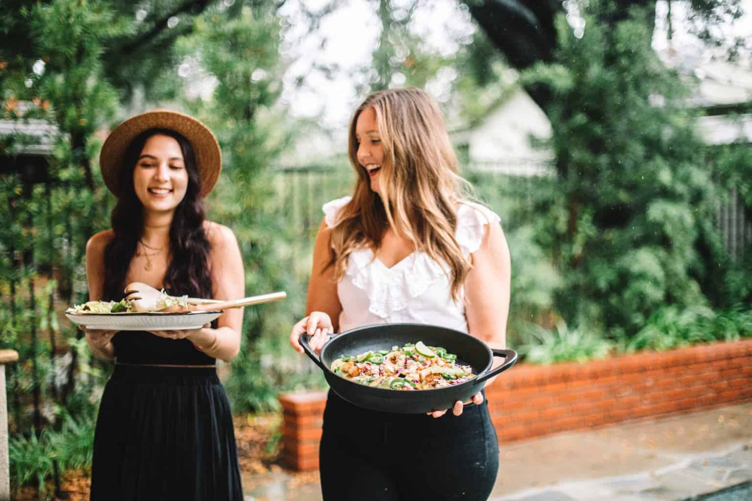 Two friends holding platters of tacos for a dinner party table.