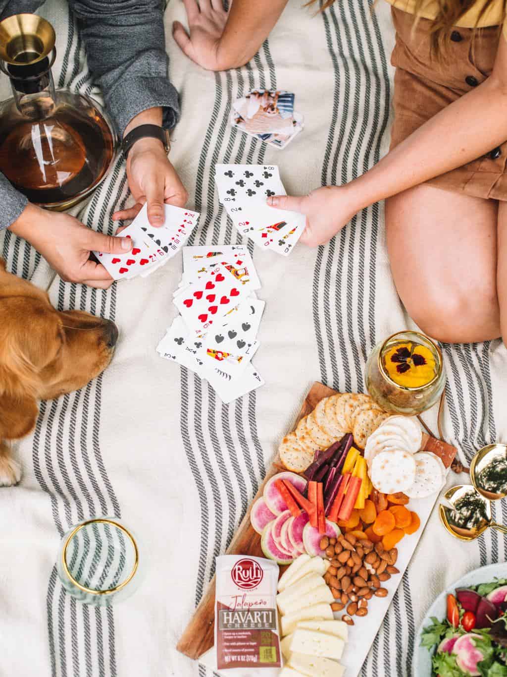 two people playing cards on a white and grey striped blanket with a golden retriever and a cheeseboard full of crackers, veggies, and havarti cheese