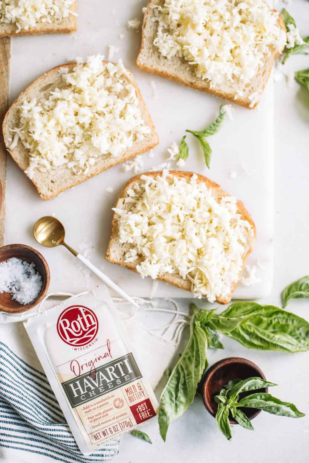 slices of bread with grated cheese on them and a package of roth havarti cheese to the side next to basil and salt