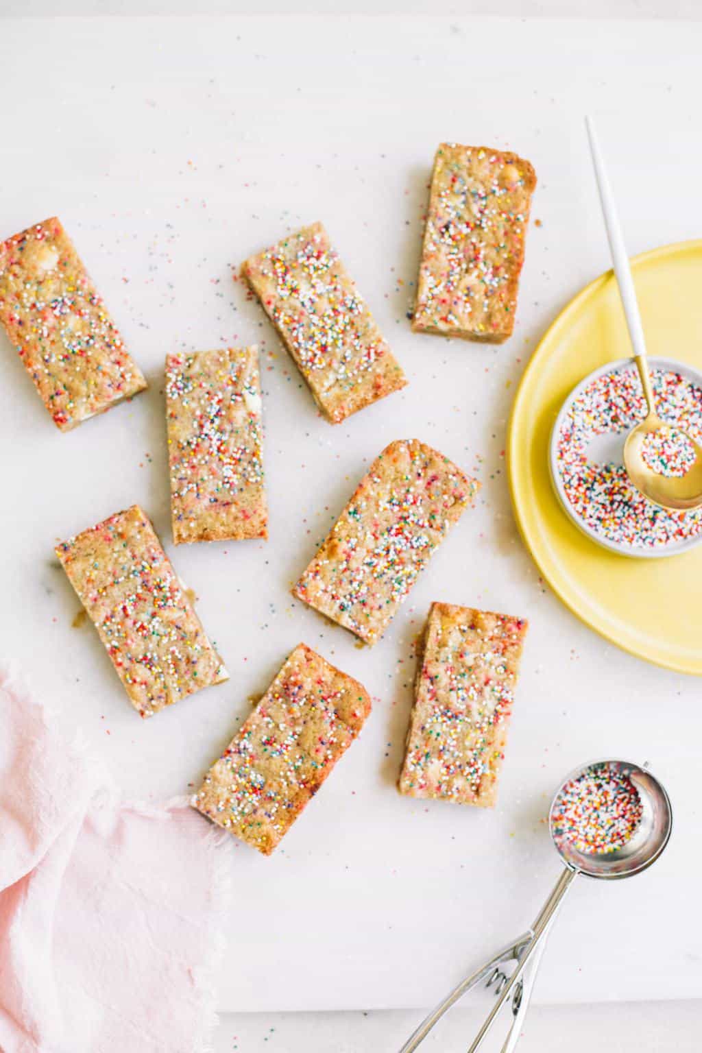 Blondies with sprinkles on a marble pastry board with yellow plate of sprinkles.