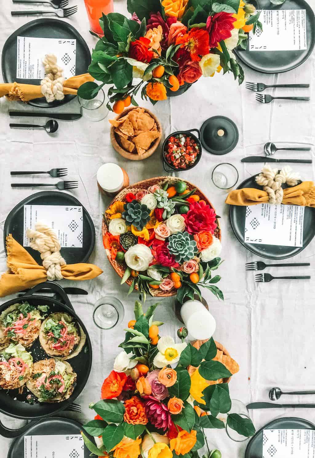 Mexican table setting with flowers, place settings and platters of tacos. 