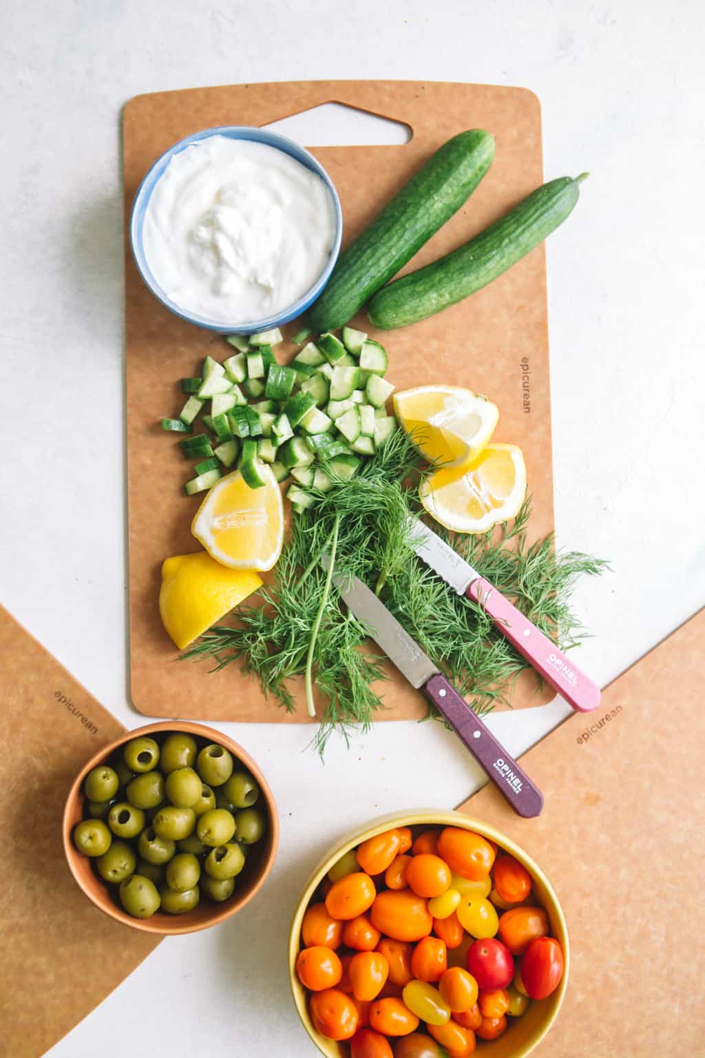 Tzatziki sauce in a bowl with chopped cucumbers, dill, and lemon wedges on a wooden board with two knives