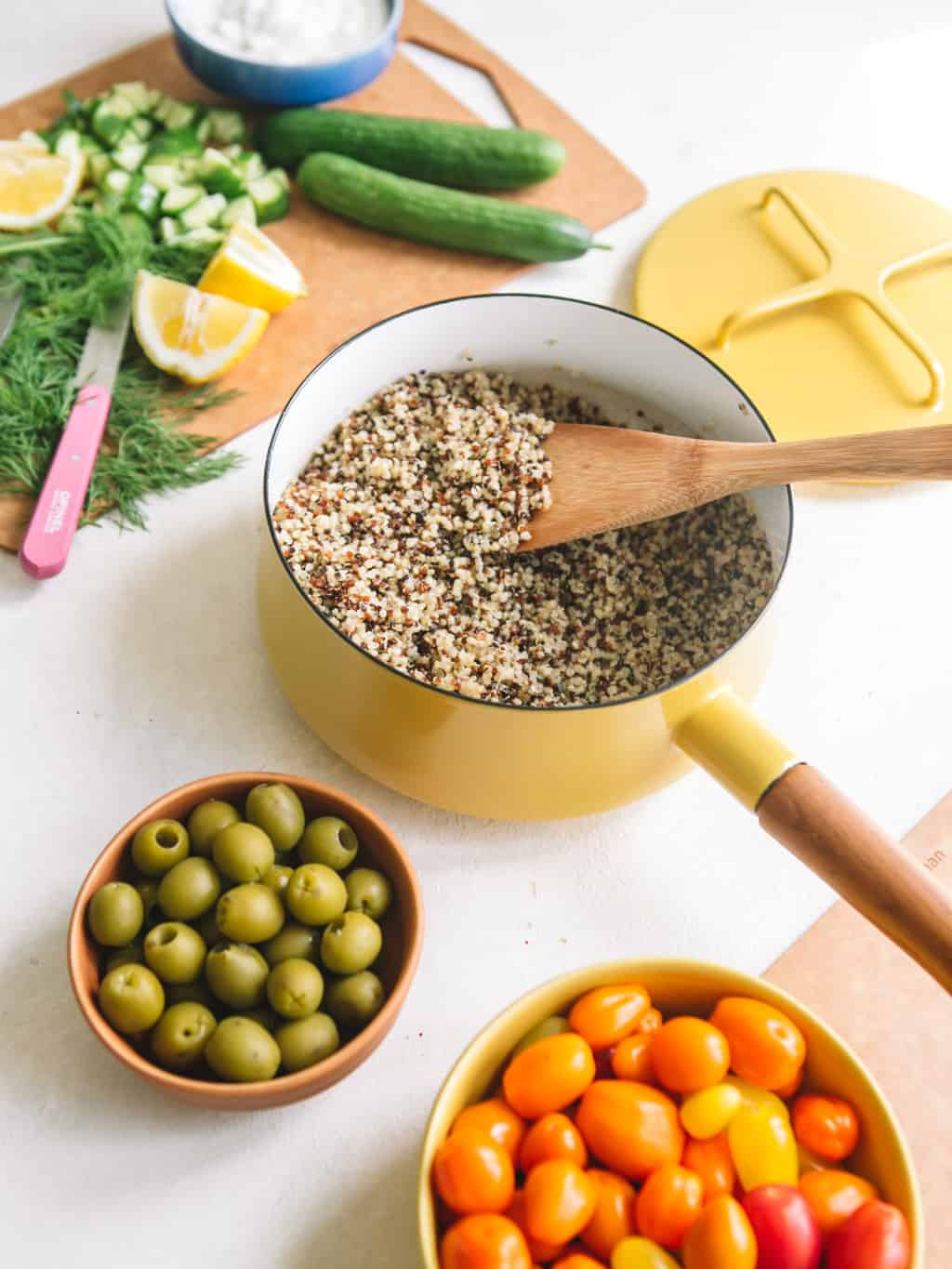 Quinoa in a yellow pot with a wooden spoon, green olives in a bowl, and cucumber and dill on a wooden board