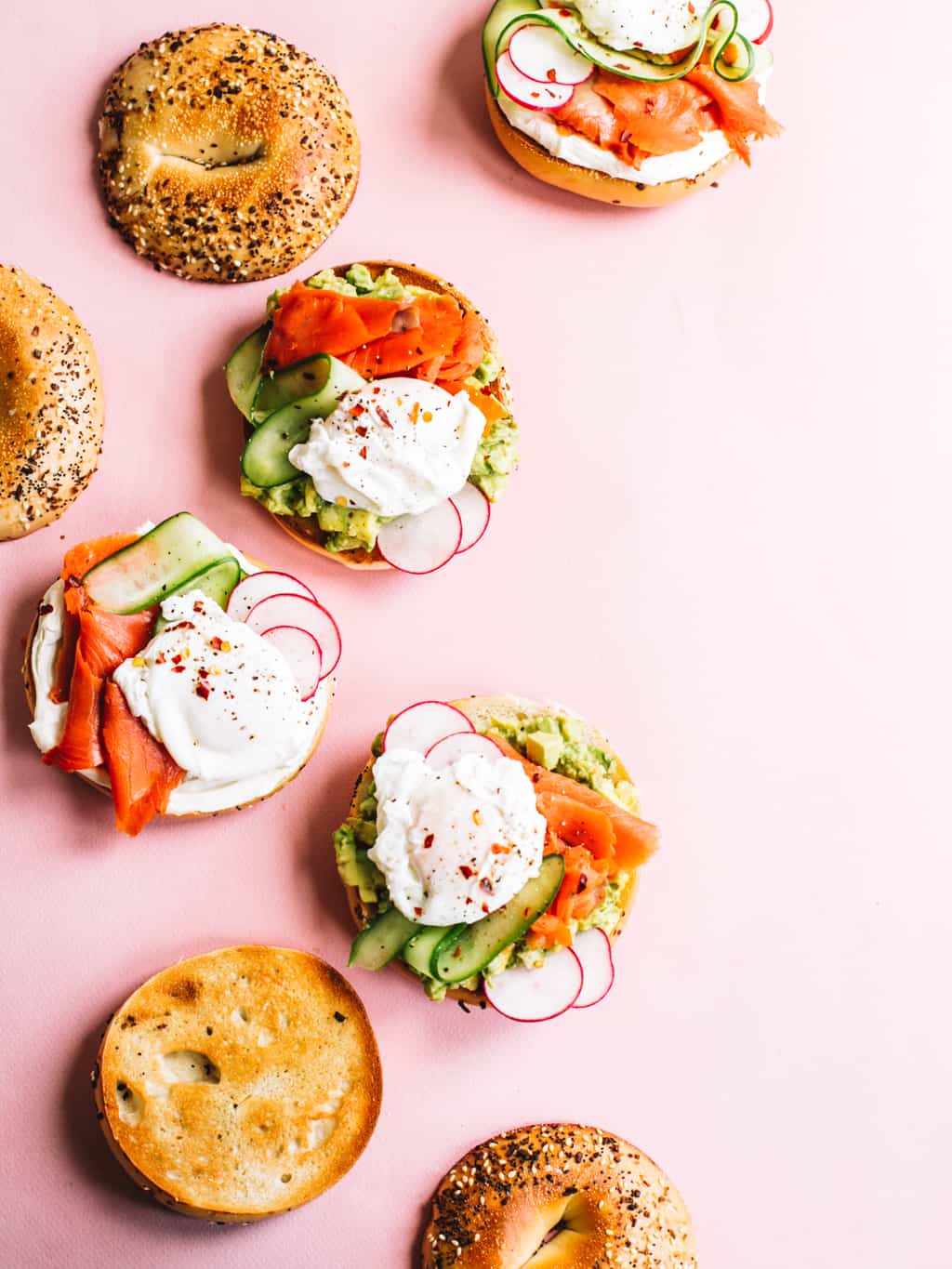 Overhead of smoked salmon bagel with mashed avocado and toppings with a pink background