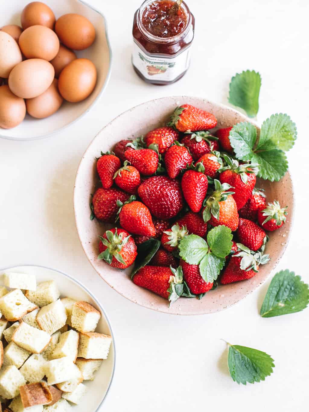 Fresh strawberries in a white bowl with bread cubes in a white bowl and eggs in a white bowl