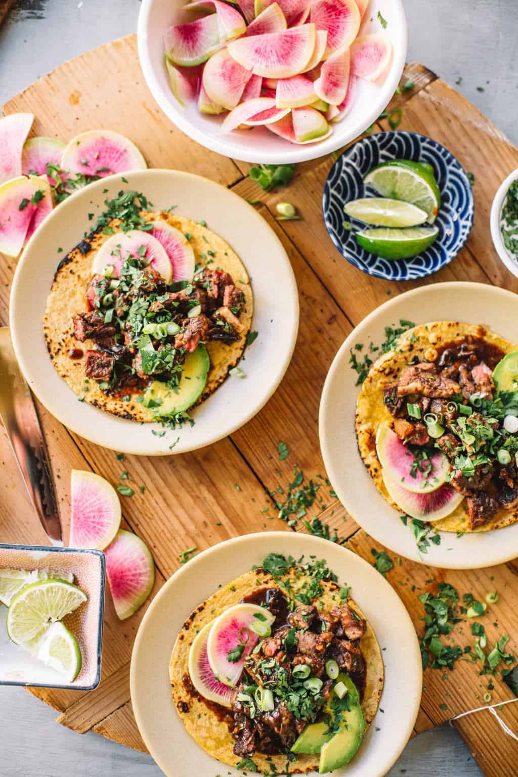 Three sous vide Asian short rib tacos on white plates with watermelon radishes and lime wedges in bowls