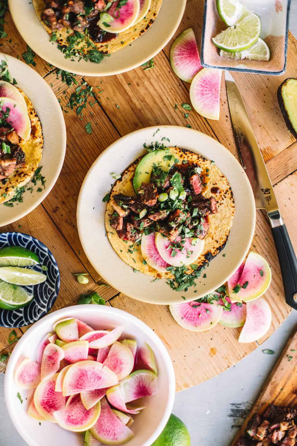 Sous vide Asian short rib tacos on a white plate with taco toppings in bowls