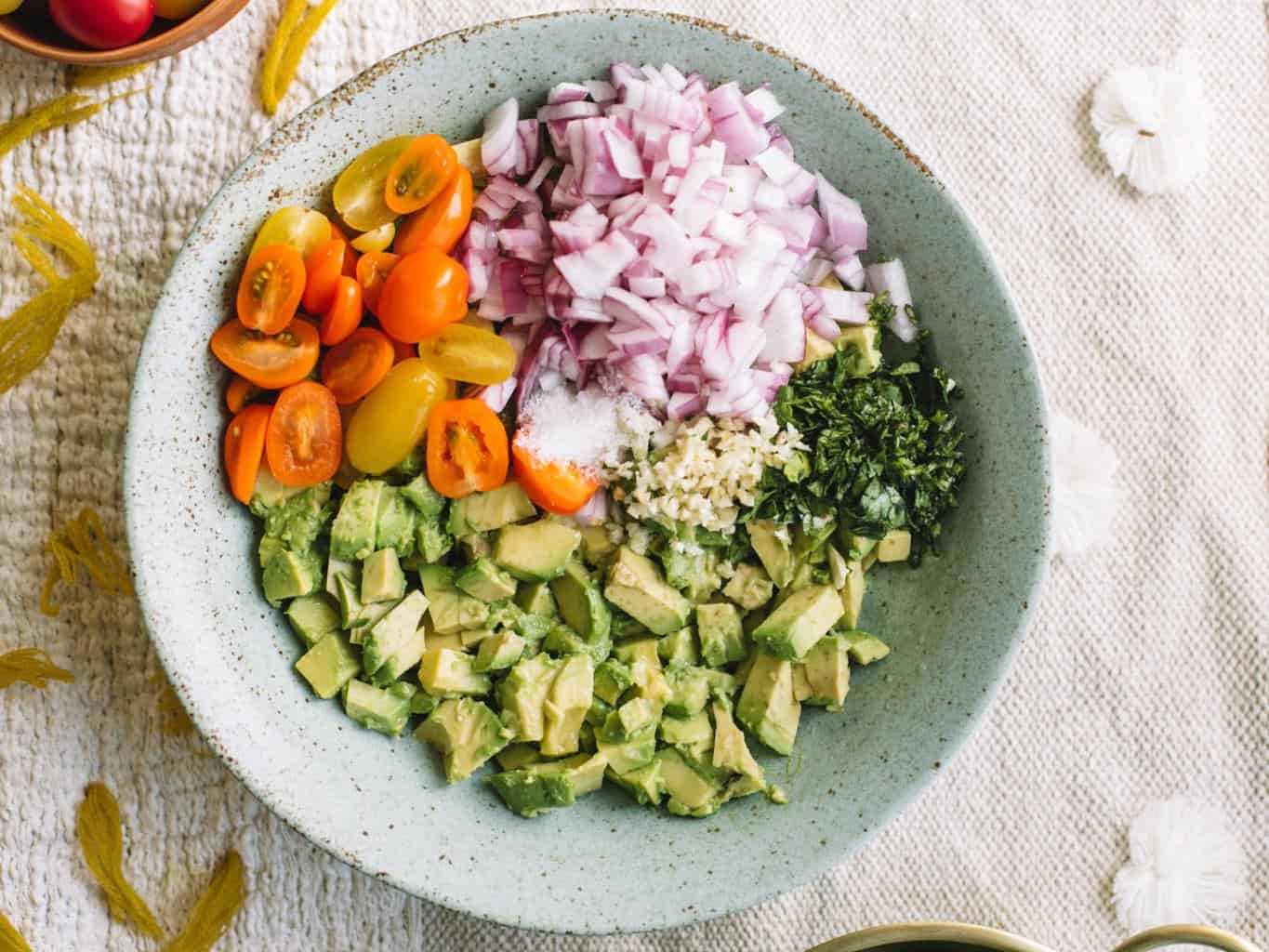 Blue bowl filled with avocado, cilantro, garlic, red onions, and cherry heirloom tomatoes.