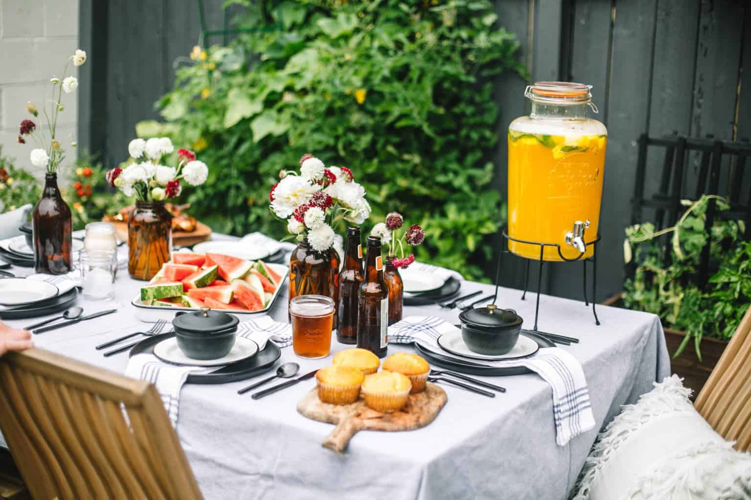 Fathers day bbq table filled with black plates, mango lemonade, watermelon platter and beer bottles full of flowers. 