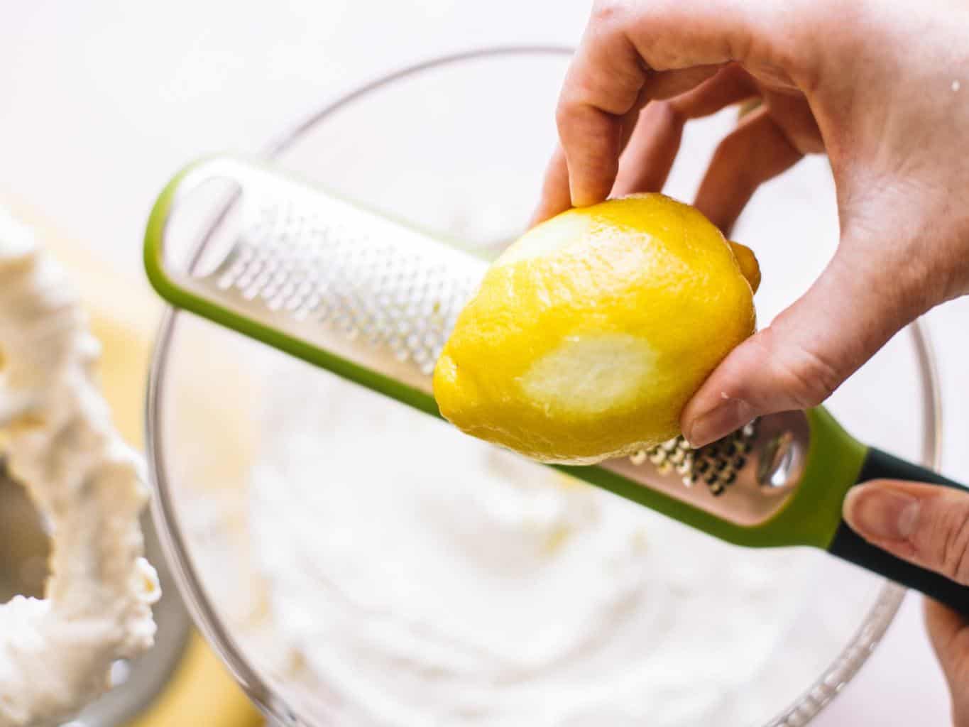 Lemon being zested over a clear bowl filled with whipped cream cheese