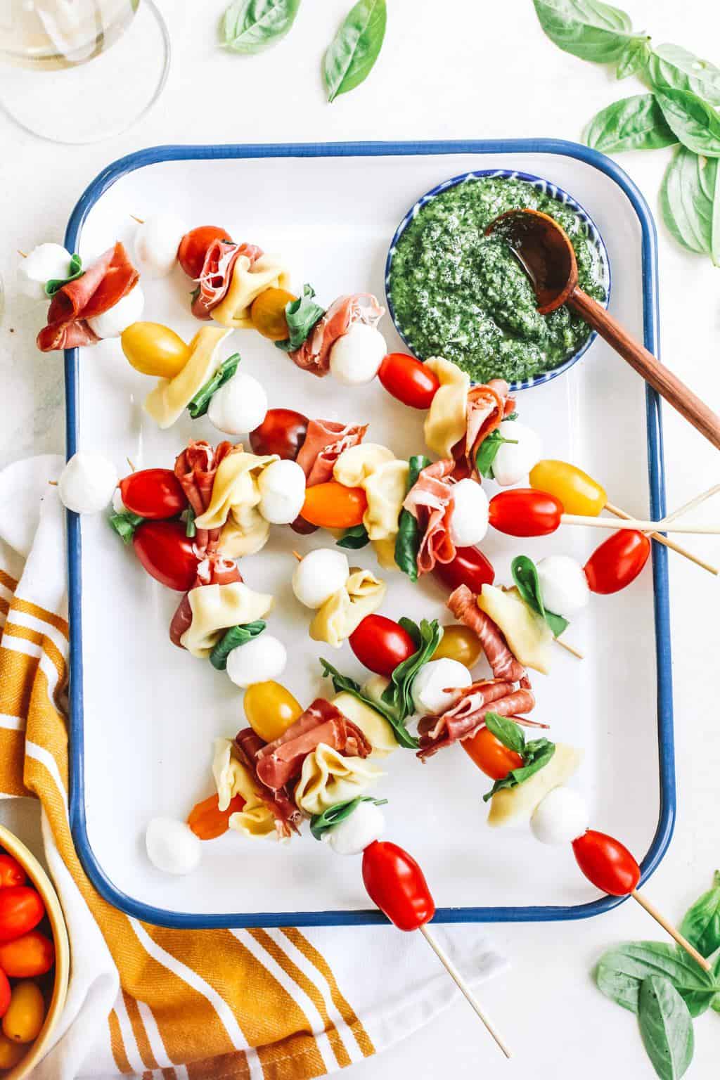 Tomato, mozzarella, basil, Prosciutto, and tortellini skewers with a bowl of pesto in white casserole dish