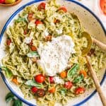 Pesto pasta salad in a white bowl with blue rim with burrata cheese and two spoons