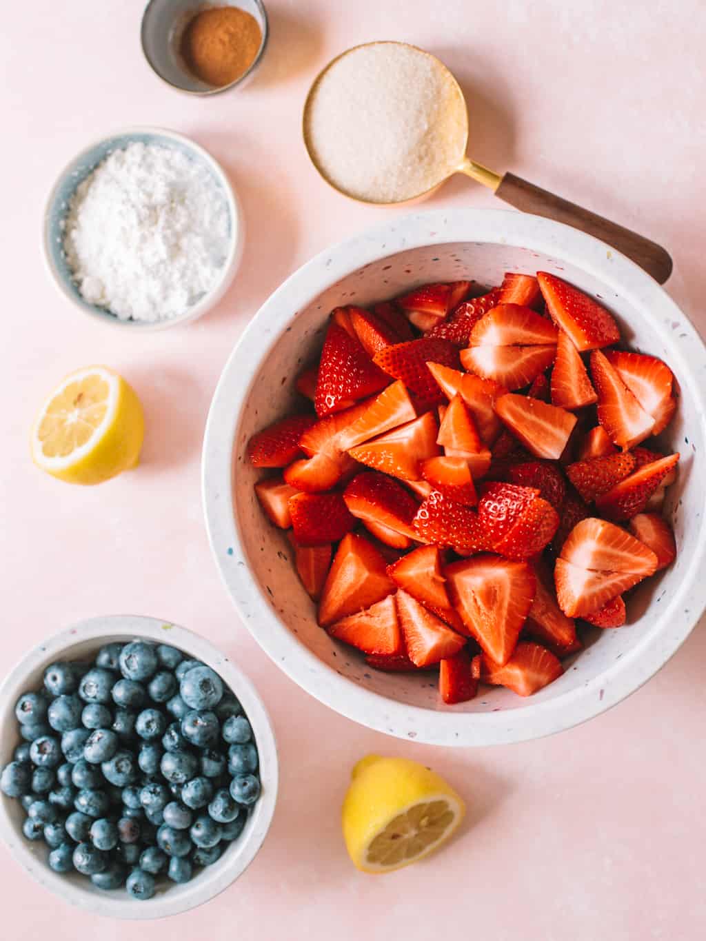 Bowl of corn starch, measuring cup of sugar, bowl of quartered strawberries and bowl of blueberries on a pink background.