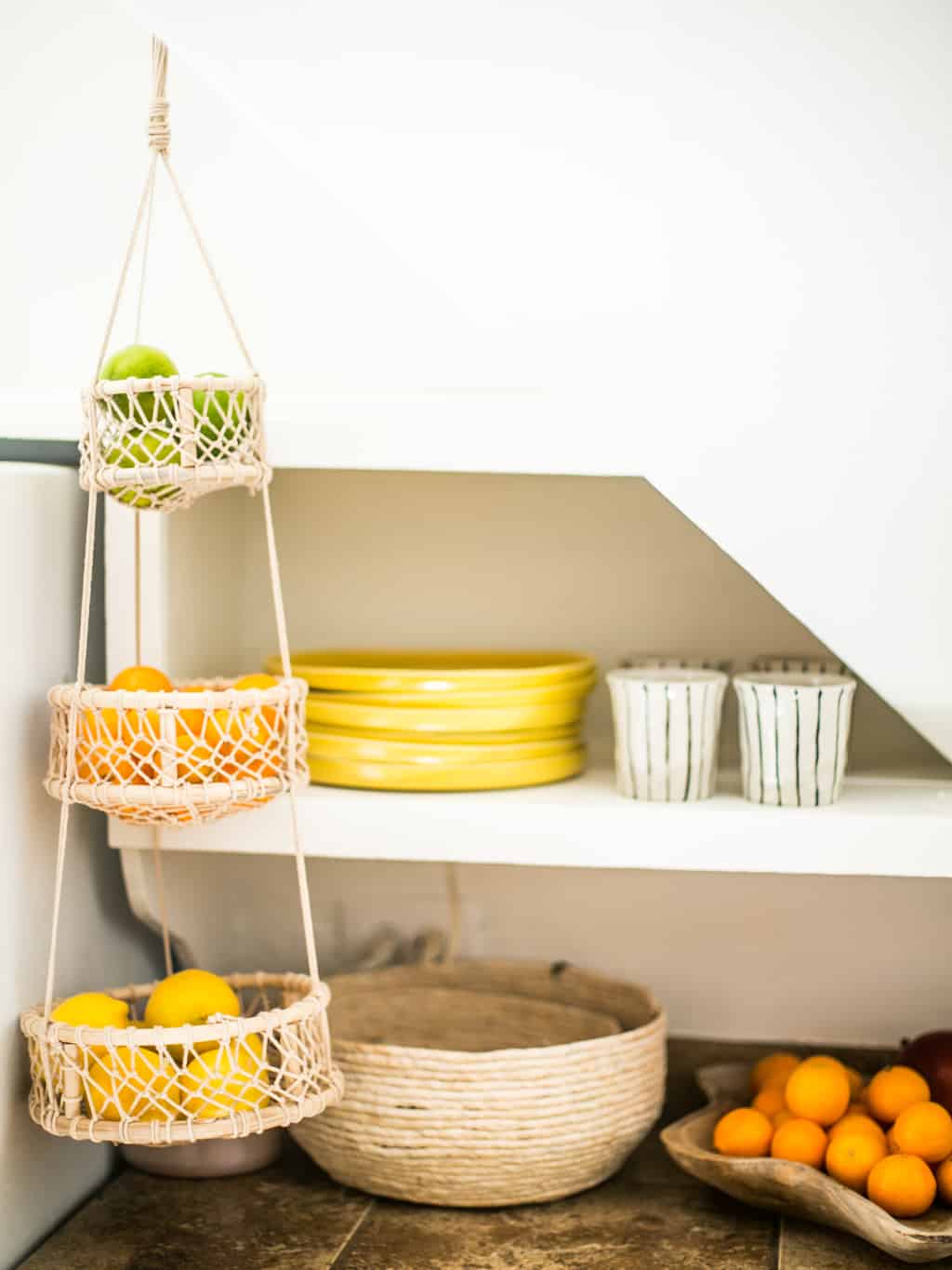 Corner of the kitchen with new yellow plates and coffee cups, with a hanging basket of fruit. 