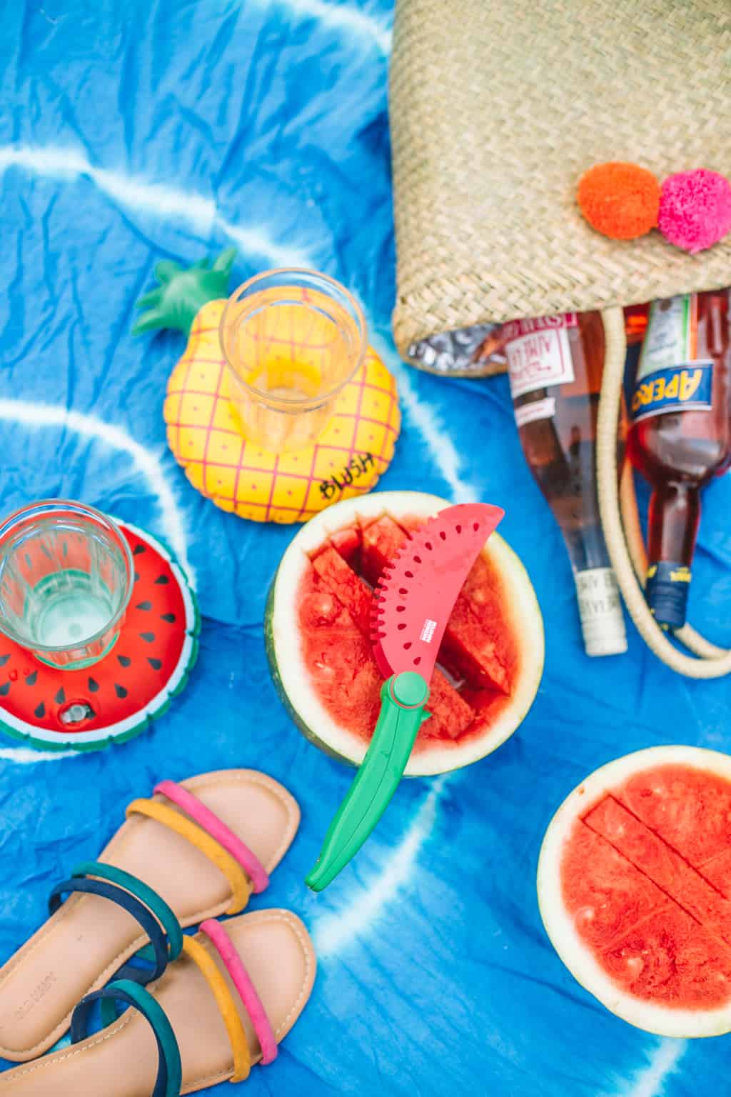 Blue tye dye beach blanket topped with sandals, watermelon and a beach bag. 